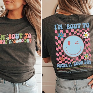 Puede incluir: Camiseta gris oscuro con la frase "I'M 'BOUT TO HAVE A GOOD DAY" en una fuente retro colorida. La parte trasera presenta una carita sonriente gui&ntilde;ando un ojo, flores y un dise&ntilde;o a cuadros rosa y negro, con texto adicional.