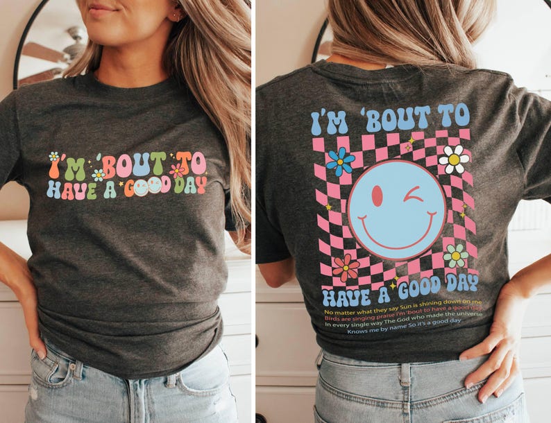 Puede incluir: Camiseta gris oscuro con la frase "I'M 'BOUT TO HAVE A GOOD DAY" en una fuente retro colorida. La parte trasera presenta una carita sonriente gui&ntilde;ando un ojo, flores y un dise&ntilde;o a cuadros rosa y negro, con texto adicional.