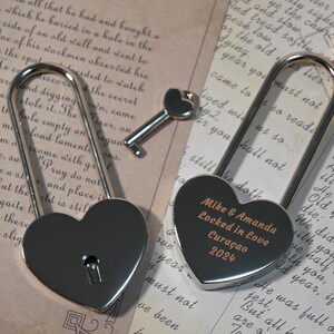 May include: Two silver heart-shaped padlocks with keys. The padlock on the right has the inscription "Mike & Amanda Locked in Love Cura&ccedil;ao 2024".
