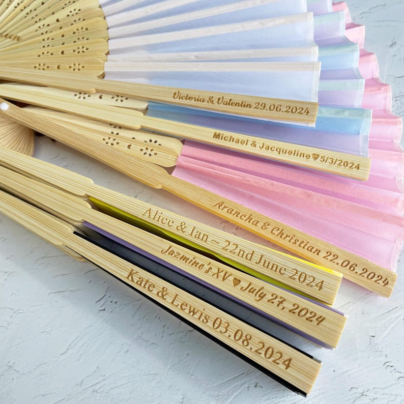 May include: A set of six bamboo hand fans with colorful silk paper and personalized inscriptions. The inscriptions include names and dates, such as "Victoria & Valentin 29.06.2024" and "Alice & Ian~22nd June 2024".