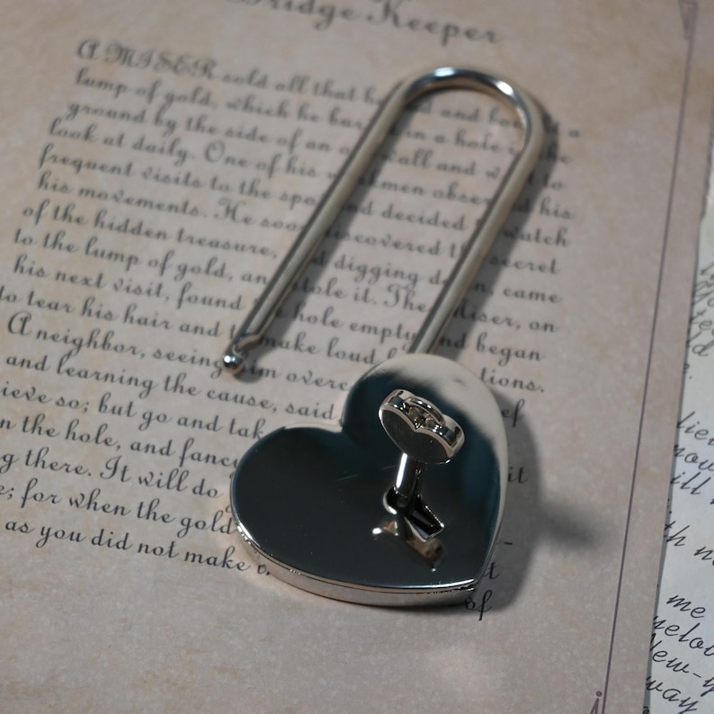 May include: A silver heart-shaped padlock with a key inserted into the keyhole. The padlock is open and the key is visible. The padlock is lying on a piece of paper with text.