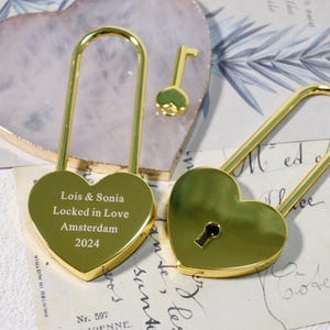 May include: Two gold heart-shaped padlocks with a key. The padlock on the left has the inscription "Lois & Sonia Locked in Love Amsterdam 2024".