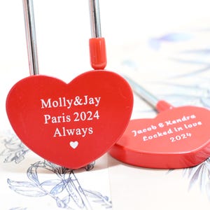 Könnte beinhalten: Zwei rote herzförmige Vorhängeschlösser mit silbernen Verschlüssen. Ein Vorhängeschloss ist mit "Molly & Jay Paris 2024 Always" graviert und das andere mit "Jacob & Kendra Locked in love 2024".