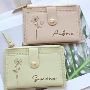 May include: Two small, rectangular wallets in beige and light green. Each wallet has a floral design and a name printed on the front. The beige wallet has the name "Aubrie" and the light green wallet has the name "Simone". Both wallets have a snap closure.