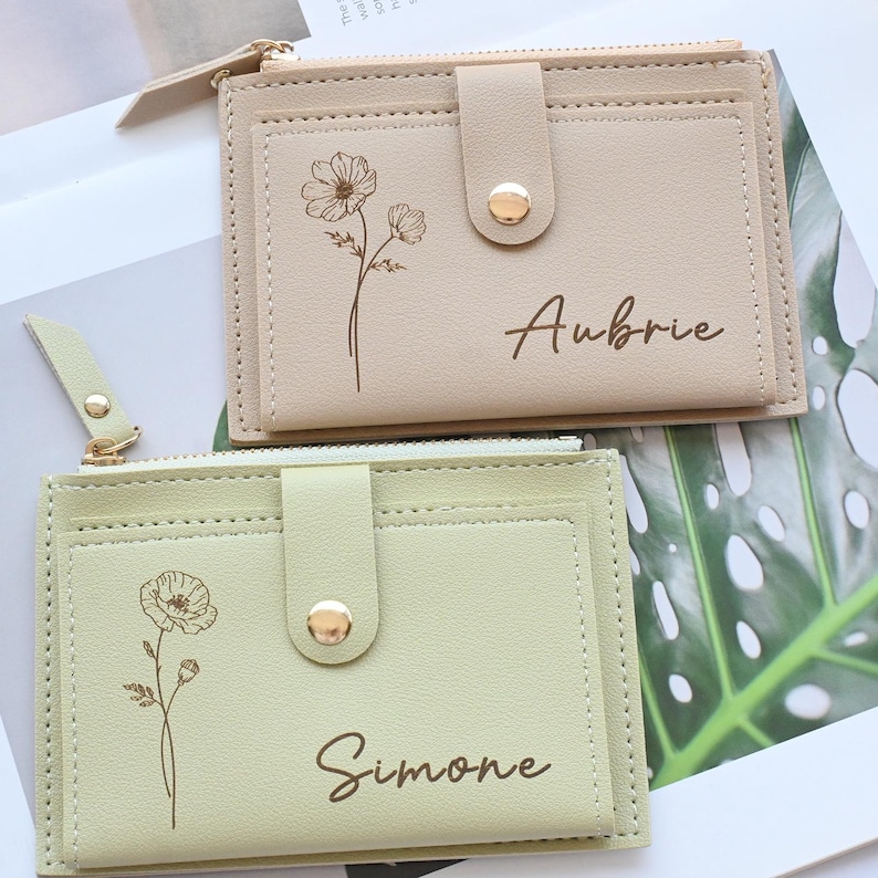 May include: Two small, rectangular wallets in beige and light green. Each wallet has a floral design and a name printed on the front. The beige wallet has the name "Aubrie" and the light green wallet has the name "Simone". Both wallets have a snap closure.