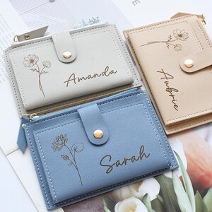 May include: Three personalized card wallets in light gray, beige, and blue. Each wallet features a floral design and a name: "Amanda", "Aubrie", and "Sarah". The wallets have a snap closure and a zipper. The wallets are made of a leather-like material.