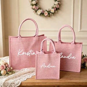 May include: Three pink tote bags of varying sizes with white lettering. The largest bag reads "Kristin", the medium bag reads "Claudia", and the smallest bag reads "Andrea". The bags have matching handles and are made of a textured material.