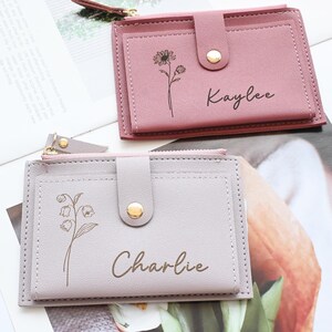 May include: Two small, rectangular wallets in shades of pink and gray. Each wallet features a floral design and a name, "Kaylee" and "Charlie", in a handwritten font. The wallets have a snap closure and a zippered compartment.