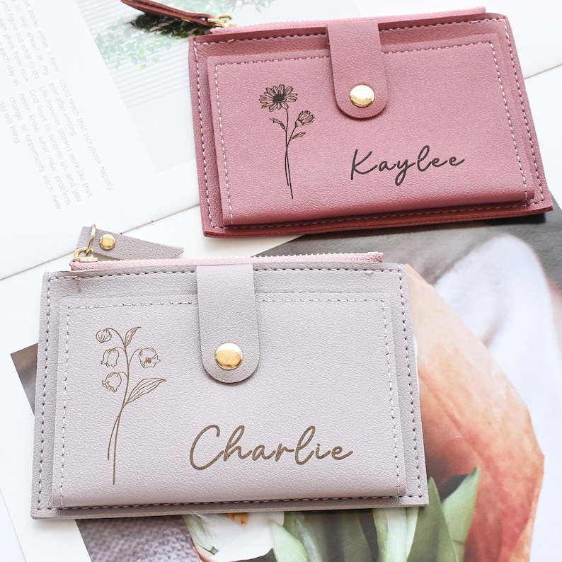 May include: Two small, rectangular wallets in shades of pink and gray. Each wallet features a floral design and a name, "Kaylee" and "Charlie", in a handwritten font. The wallets have a snap closure and a zippered compartment.