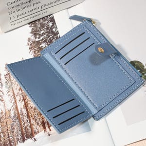 May include: Open, light blue wallet with multiple card slots and a zippered compartment. The wallet is made of a smooth material and features a snap closure with a gold-colored button. The text "Cont 100% Ecog Ne le jetez pas. I 1 peut servir plusieurs No one can replace you" is visible.