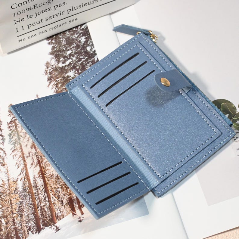 May include: Open, light blue wallet with multiple card slots and a zippered compartment. The wallet is made of a smooth material and features a snap closure with a gold-colored button. The text "Cont 100% Ecog Ne le jetez pas. I 1 peut servir plusieurs No one can replace you" is visible.