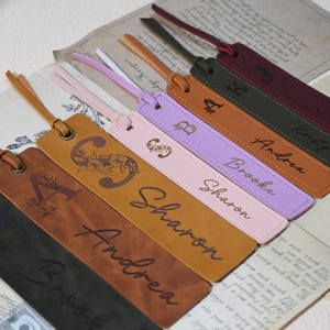 May include: A set of eight personalised leather bookmarks with tassels. The bookmarks are brown, pink, purple, and green. Each bookmark has a name and a letter engraved on it. The names are Andrea, Brooke, Sharon, and Kristy.