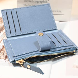 May include: Open light blue wallet with multiple card slots and a zippered compartment. The wallet has a snap closure and gold-colored zipper and hardware. The stitching is white, and the wallet is made of a smooth material.