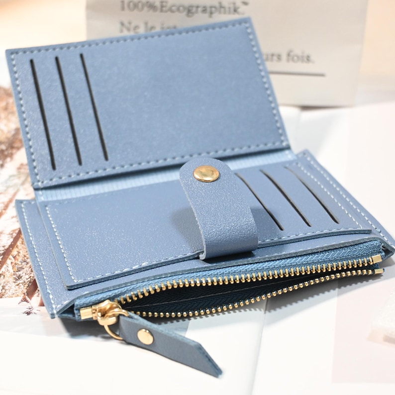 May include: Open light blue wallet with multiple card slots and a zippered compartment. The wallet has a snap closure and gold-colored zipper and hardware. The stitching is white, and the wallet is made of a smooth material.