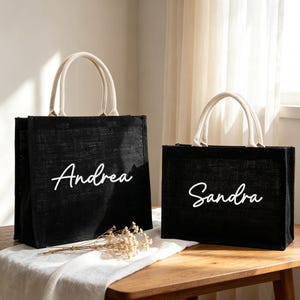 Könnte beinhalten: Zwei schwarze, rechteckige Tragetaschen aus strukturiertem Webmaterial. Jede Tasche verfügt über zwei cremefarbene Stoffgriffe und den Namen Andrea oder Sandra, der in weißer Schreibschrift auf der Vorderseite der jeweiligen Tasche aufgedruckt ist.