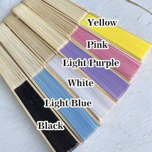 May include: A set of seven hand fans in various colors, including black, light blue, white, light purple, pink, yellow, and natural wood. The fans are arranged in a row, with the black fan at the bottom and the natural wood fan at the top.