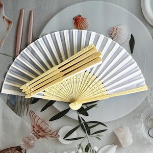 May include: A set of white and gray paper fans with wooden handles. The handles are engraved with the text "Phoebe & Owen 08.09.2024".