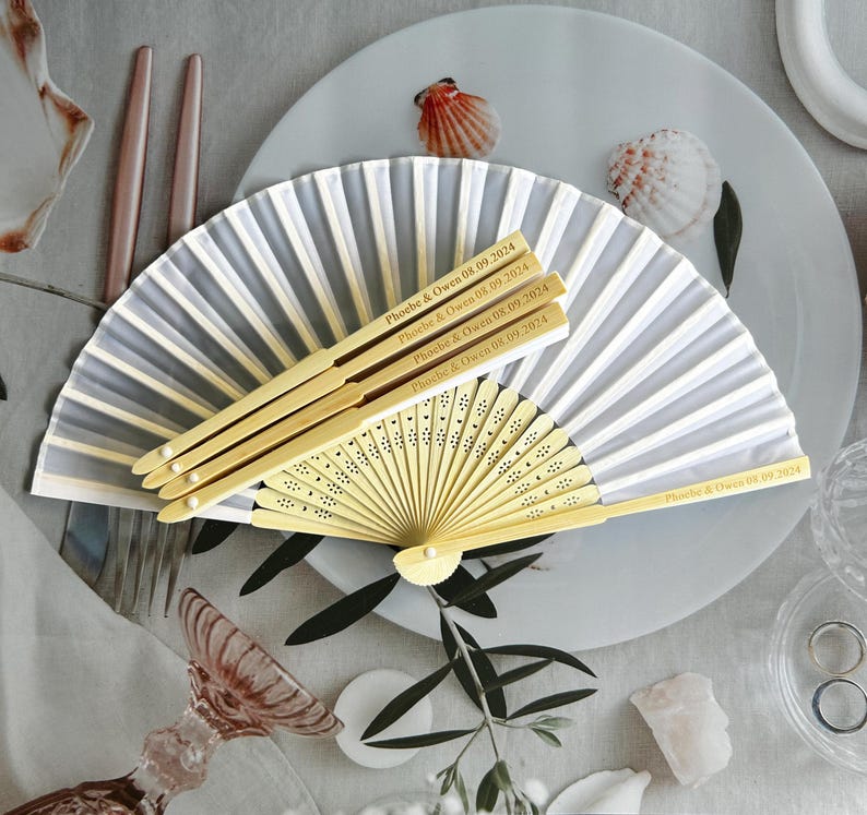 May include: A set of white and gray paper fans with wooden handles. The handles are engraved with the text "Phoebe & Owen 08.09.2024".