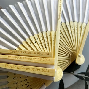 May include: A set of white paper folding fans with bamboo handles. The handles are engraved with the text "Phoebe & Owen 08.09.2024".