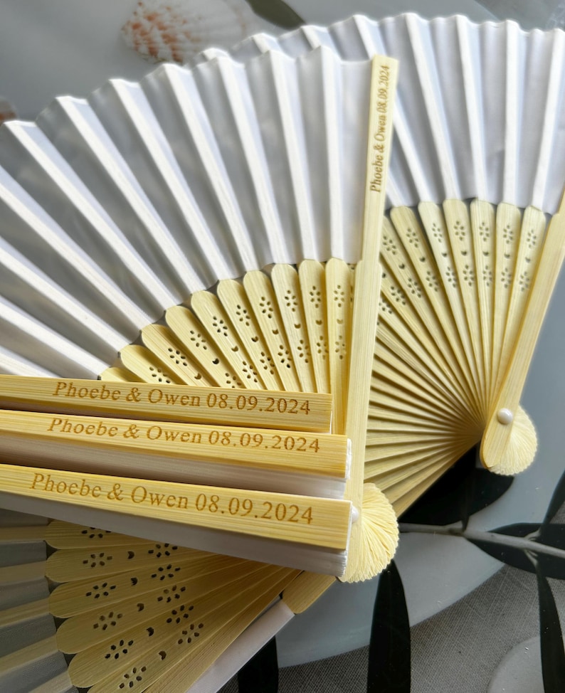 May include: A set of white paper folding fans with bamboo handles. The handles are engraved with the text "Phoebe & Owen 08.09.2024".