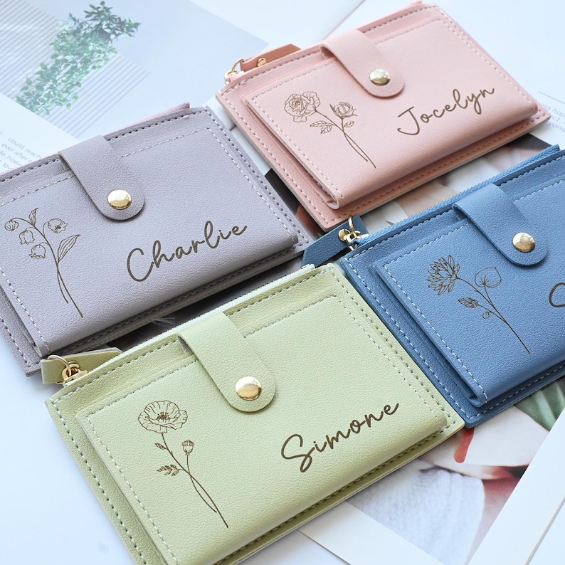 May include: A collection of personalized cardholders in pastel colors: lavender, pink, blue, and green. Each wallet features a floral design and a name, such as "Charlie" or "Simone". The wallets have a snap closure and a zippered pocket.