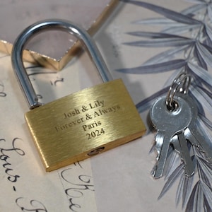 May include: A gold padlock with a silver shackle and two keys. The padlock has the inscription "Josh & Lily Forever & Always Paris 2024" engraved on it.