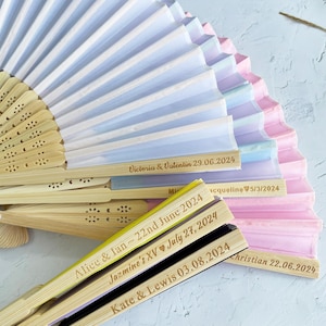 May include: A stack of colorful paper fans with wooden handles. Each fan has a personalized message with a name and date, such as "Victoria & Valentin 29.06.2024" and "Alice & Ian ~ 22nd June 2024".