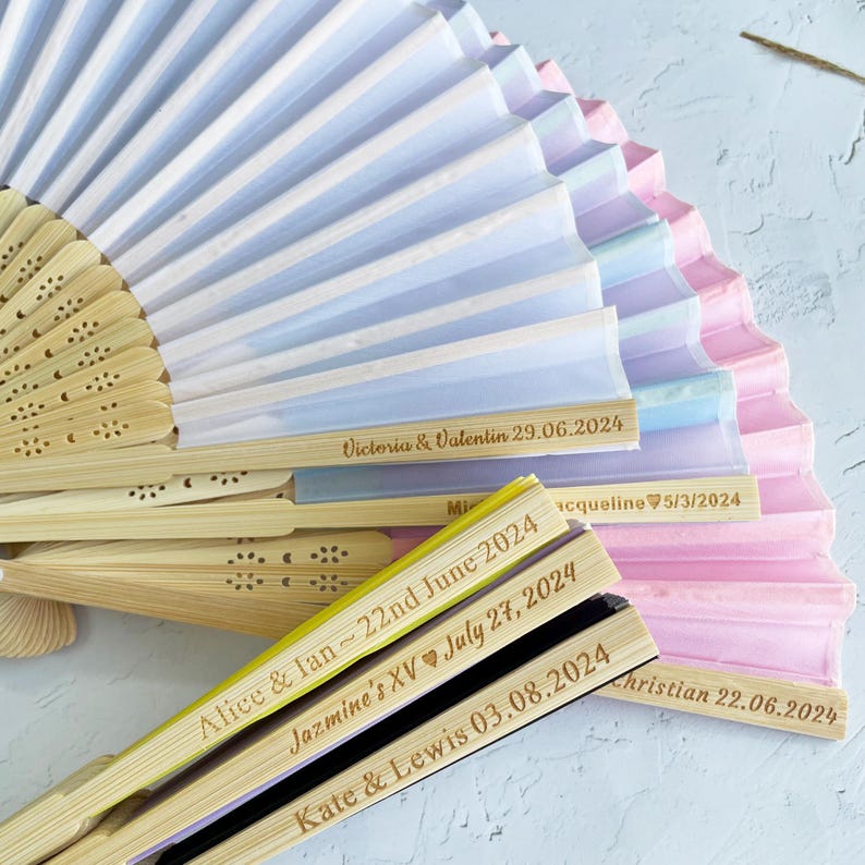 May include: A stack of colorful paper fans with wooden handles. Each fan has a personalized message with a name and date, such as "Victoria & Valentin 29.06.2024" and "Alice & Ian ~ 22nd June 2024".
