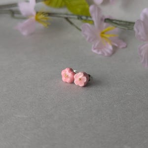 以下が含まれることがあります： 小さなピンクの桜の花の形をしたスタッドピアス一対。各ピアスは5枚の丸みを帯びた花びらで構成され、中央に小さな金色の点が施されています。耳たぶに装着するための銀色の金属製ポストが付いています。