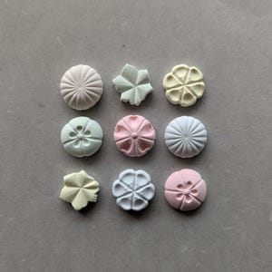 Könnte beinhalten: Eine Ansammlung von neun kleinen, mattierten Dekorationsobjekten in Form von Blumen und kreisförmigen Medaillons in Pastelltönen wie Rosa, Gelb, Blau, Grün und Weiß, angeordnet in einem drei-mal-drei Raster auf einer neutralen grauen Fläche.