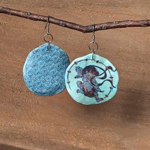 Op de afbeelding: Een paar blauwe en groene keramische oorbellen met een draaiend patroon en een Japans geïnspireerd ontwerp. De ene oorbel heeft een blauw en wit draaiend patroon, terwijl de andere oorbel een groene achtergrond heeft met een zwart-witte afbeelding van een figuur in traditionele Japanse kleding.