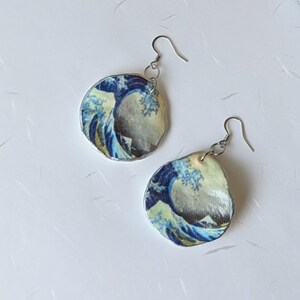 May include: A pair of dangle earrings featuring a print of Hokusai's "The Great Wave off Kanagawa." The earrings have a silver-colored hook and a circular, irregular shape. The wave design includes shades of blue, white, and yellow.