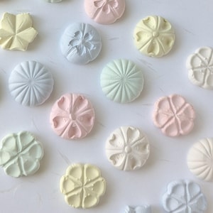 May include: Assortment of pastel-coloured ceramic flower-shaped buttons. The buttons are in various shades of blue, pink, green, and yellow.