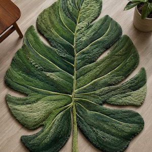 May include: A green leaf-shaped rug, resembling a Monstera plant leaf, with detailed veining and a textured surface. The rug's varying shades of green create a realistic design. It is placed on a light wooden floor, adding a touch of nature to the room.