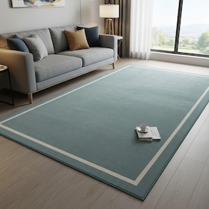 Könnte beinhalten: Ein rechteckiger, türkisfarbener Teppich mit cremefarbenem Rand, auf einem Holzboden. Der Teppich befindet sich in einem Wohnzimmer, mit einem grauen Sofa und Kissen im Hintergrund. Eine Kaffeetasse und ein Buch liegen auf dem Teppich.