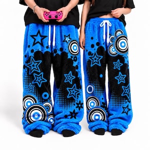 May include: Two pairs of bright blue fuzzy fleece lounge trousers featuring a black graphic print of stars, concentric circles, and dotted patterns. Each pair has a white drawstring at the elastic waistband and a loose, relaxed fit design.