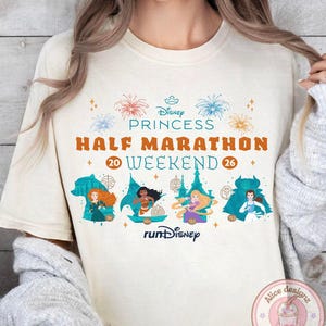May include: Cream-colored t-shirt featuring the text "Disney Princess Half Marathon Weekend 26" with illustrations of Disney princesses. The design includes fireworks and the "runDisney" logo. The shirt has short sleeves.