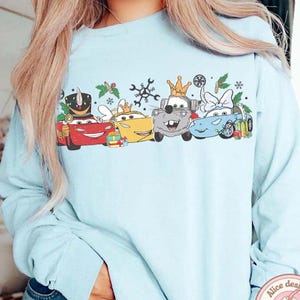 May include: Light blue long-sleeve sweatshirt featuring a festive design of cartoon cars with crowns and holiday decorations. The cars are red, yellow, gray, and blue. The design includes snowflakes and sprigs of holly.