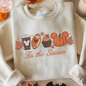 May include: Cream-colored sweatshirt with an embroidered fall-themed design. The design includes a coffee cup, football, cupcake, and pumpkin, all with Mickey Mouse-inspired details. The text "Tis the Season" is below the design. A small pumpkin and leaves are visible.
