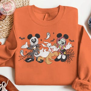 May include: Orange sweatshirt featuring a Halloween-themed graphic with cartoon characters. The design includes Mickey Mouse dressed as Jack Skellington, Pluto as a ghost, and Minnie Mouse as a zombie, with bats and spiderwebs.