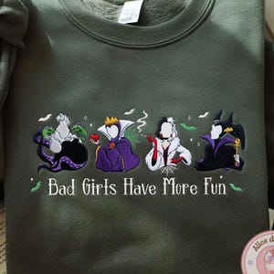 May include: An olive green sweatshirt featuring embroidered cartoon villains. The design includes Ursula, the Evil Queen, Cruella de Vil, and Maleficent. The text "Bad Girls Have More Fun" is also embroidered on the sweatshirt.