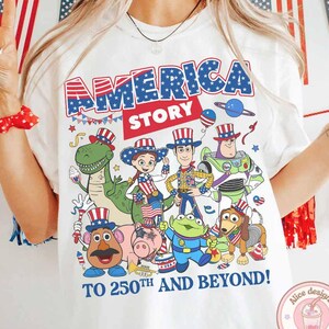 May include: White t-shirt with the text "AMERICA STORY" in blue and red, featuring cartoon characters in patriotic attire. The phrase "TO 250TH AND BEYOND!" is printed below the characters. The shirt has a round neck.