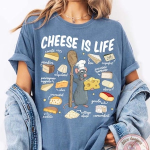 Puede incluir: Una camiseta azul con el texto "CHEESE IS LIFE" e ilustraciones de varios quesos. Una rata de dibujos animados con un gorro de chef sostiene una cuchara de madera y una cu&ntilde;a de queso. La camiseta tambi&eacute;n presenta nombres de quesos.