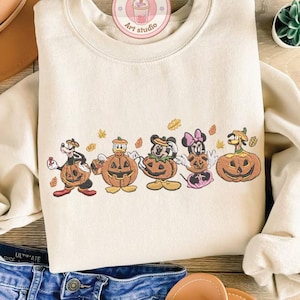 Puede incluir: Sudadera de color crema con un dise&ntilde;o bordado de personajes de dibujos animados disfrazados de calabazas. El dise&ntilde;o incluye un perro, un pato, Mickey Mouse, Minnie Mouse y un personaje con cabeza de calabaza. La sudadera tiene cuello redondo y mangas largas.