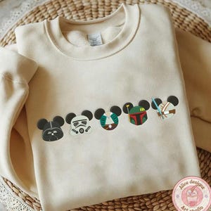 May include: Cream-colored sweatshirt featuring embroidered character designs. The designs include Darth Vader, a Stormtrooper, and other characters. The sweatshirt has a crew neck and long sleeves. The design is a mashup of Disney and Star Wars.