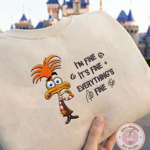 May include: Beige sweatshirt featuring an embroidered cartoon character with orange hair and a brown outfit. The text "I'M FINE, IT'S FINE, EVERYTHING'S FINE" is printed on the sweatshirt.
