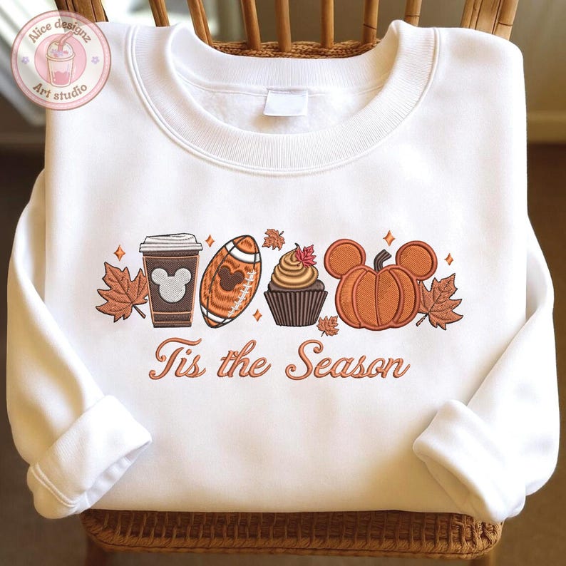 May include: White sweatshirt with an embroidered fall-themed design. The design includes a coffee cup, football, cupcake, and pumpkin, all in shades of brown and orange. The text "Tis the Season" is below the design.
