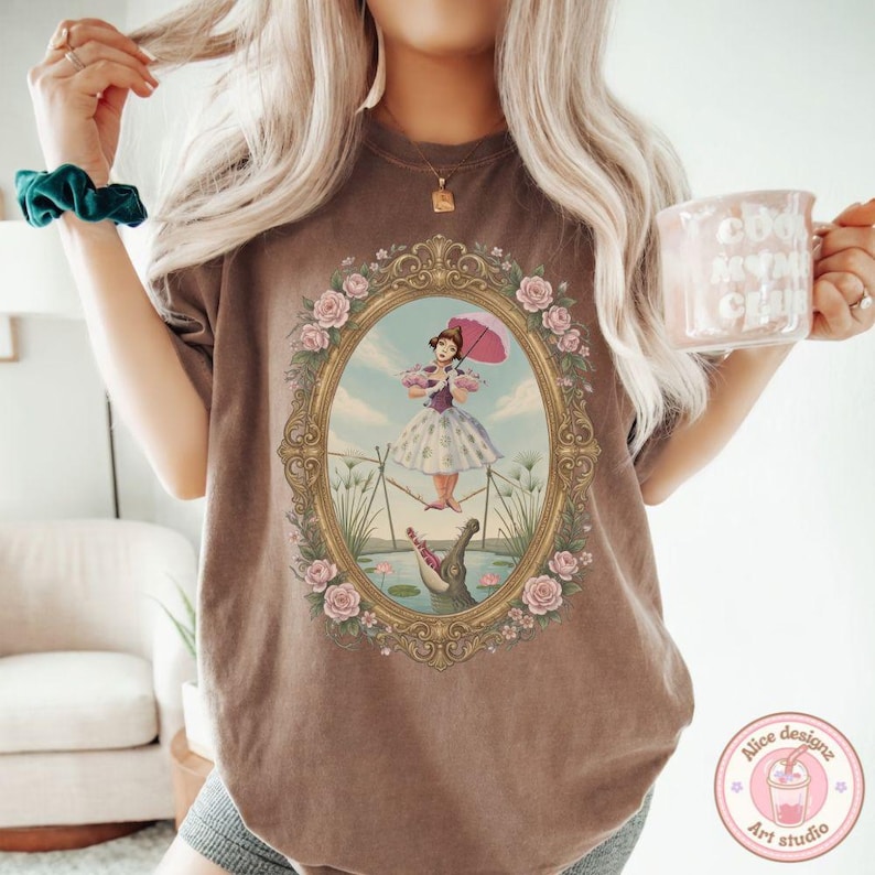 May include: A brown t-shirt featuring a graphic print of a person in a dress walking a tightrope above an alligator, enclosed in an ornate gold oval frame decorated with pink roses. The design includes a pink umbrella and a blue sky background.
