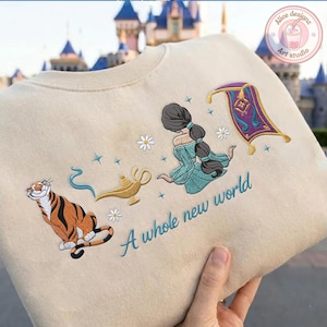 May include: A beige sweatshirt featuring embroidered designs of a tiger, a gold lamp with blue smoke, a person with a long dark braid in a teal dress, a purple magic carpet, and white daisies, with the text A whole new world in teal script.