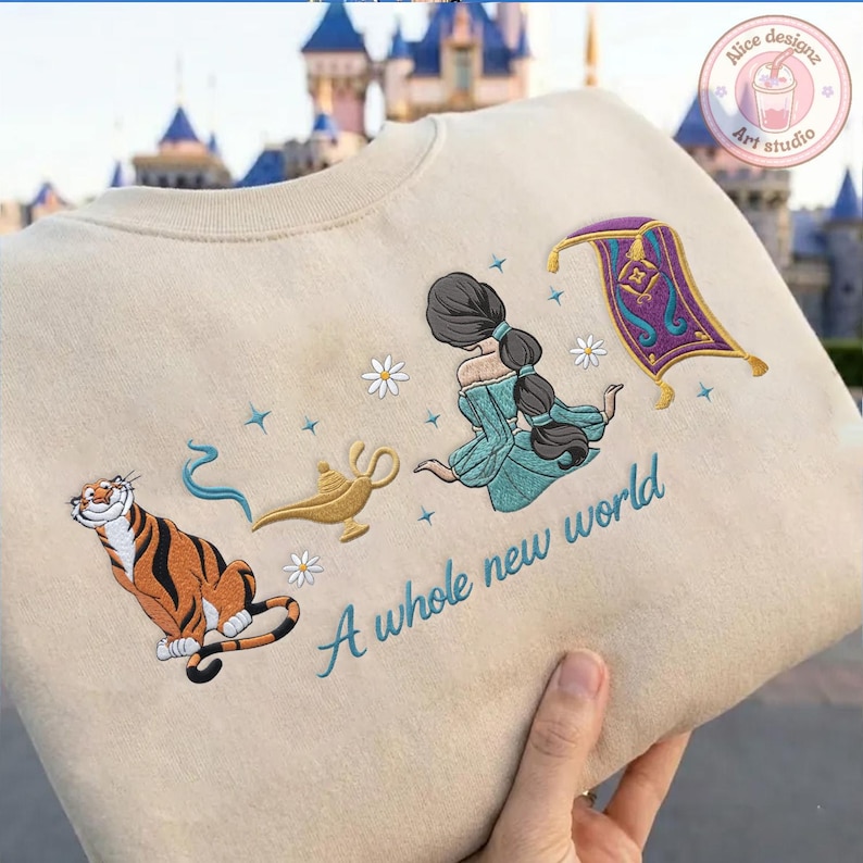 May include: A beige sweatshirt featuring embroidered designs of a tiger, a gold lamp with blue smoke, a person with a long dark braid in a teal dress, a purple magic carpet, and white daisies, with the text A whole new world in teal script.